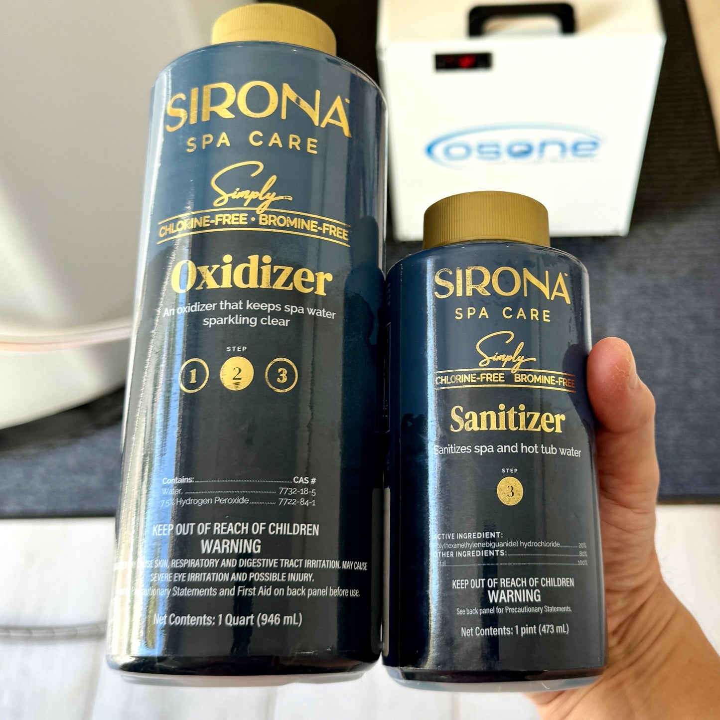 Two bottles of Sirona Spa Care chemicals, Oxidizer and Sanitizer, held by a hand. The bottles are dark blue with gold caps and gold lettering, with a white Osone device in the background.